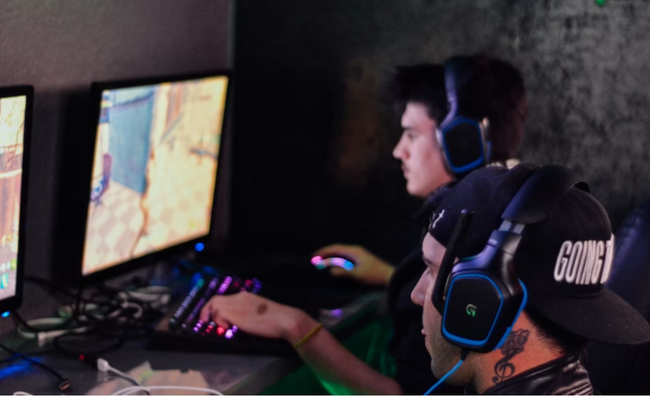 ALT: Duas pessoas jogando videogame em computadores em um ambiente de gaming, usando fones de ouvido